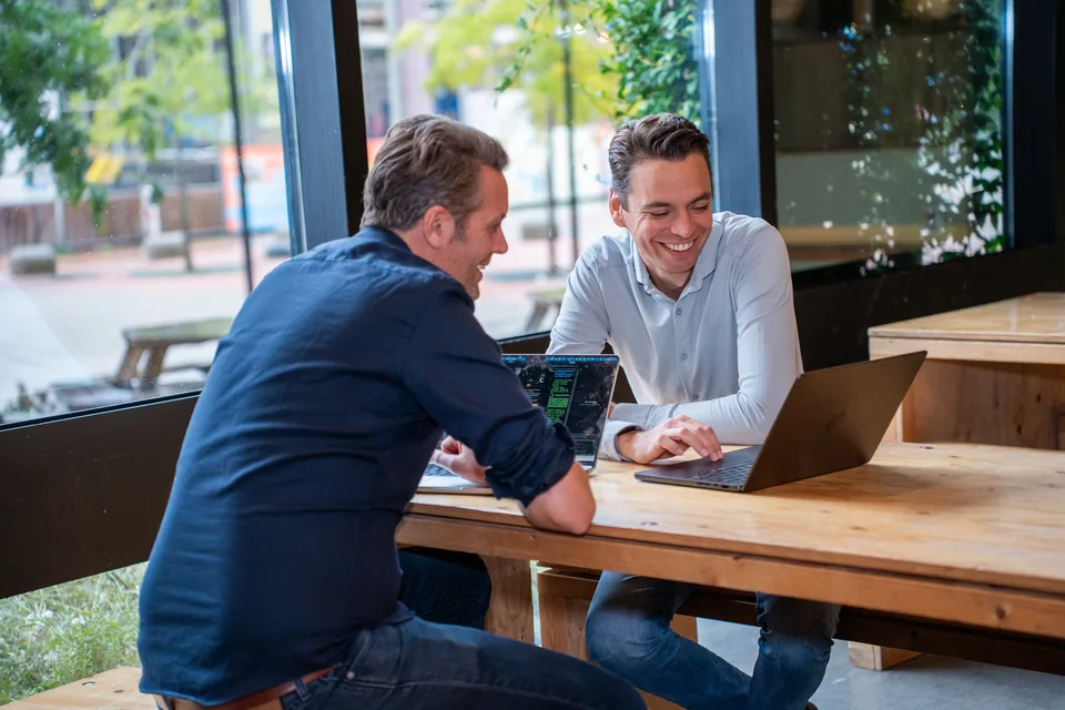 Twee ondernemers overleggen lachend aan tafel met hun laptops