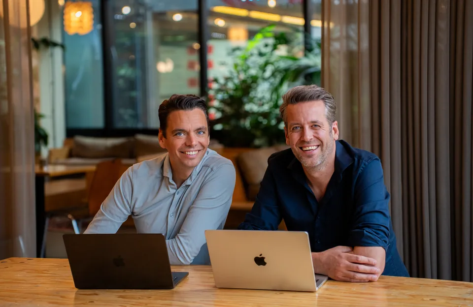 Twee ondernemers achter hun laptops kijken lachend in de camera