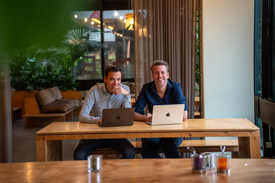 Twee ondernemers aan een lange houten tafel met hun laptops
