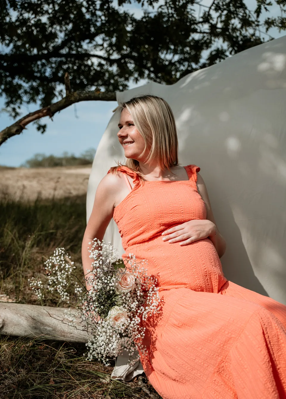 Zwangere vrouw in een oranje jurk zit op een omgevallen boom